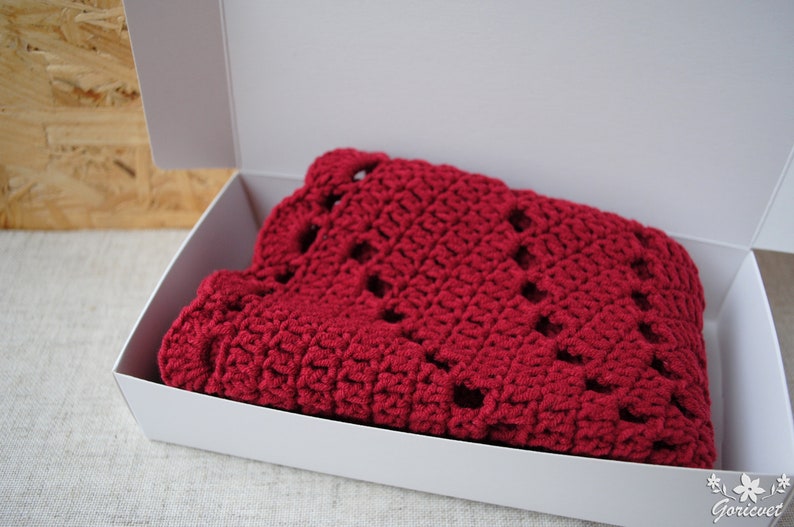 Pu&ograve; includere: Una coperta di uncinetto rossa piegata in una scatola bianca. La coperta ha un motivo a pizzo.