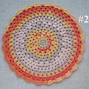 Puede incluir: Un tapete de ganchillo circular hecho a mano en tonos naranja, amarillo y gris. El tapete presenta un borde festoneado y un dise&ntilde;o central con el n&uacute;mero "#2" en blanco. Es un art&iacute;culo decorativo.