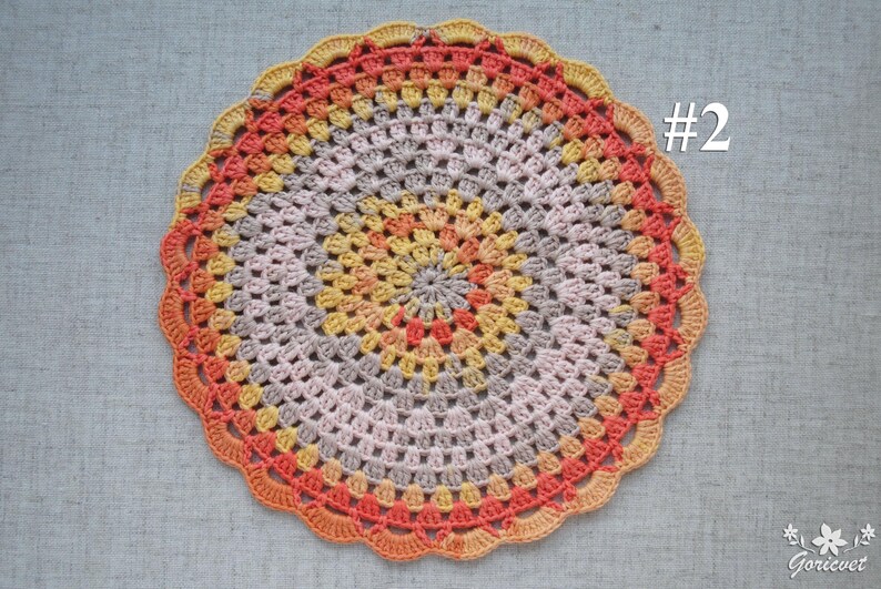 Puede incluir: Un tapete de ganchillo circular hecho a mano en tonos naranja, amarillo y gris. El tapete presenta un borde festoneado y un dise&ntilde;o central con el n&uacute;mero "#2" en blanco. Es un art&iacute;culo decorativo.