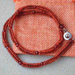 May include: A handcrafted necklace featuring multiple strands of small, reddish-brown beads. The necklace is secured with a silver-colored clasp and rests on a textured orange fabric. The beads are arranged in a braided pattern.