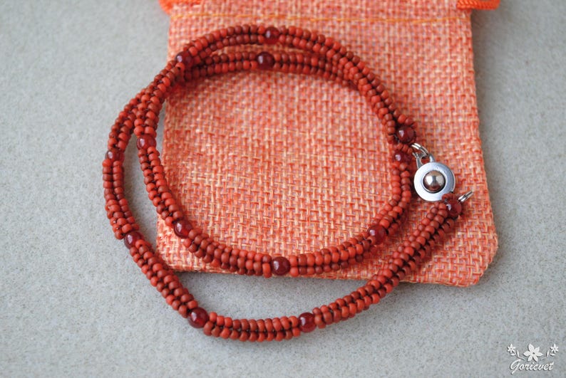 May include: A handcrafted necklace featuring multiple strands of small, reddish-brown beads. The necklace is secured with a silver-colored clasp and rests on a textured orange fabric. The beads are arranged in a braided pattern.