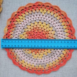 Puede incluir: Un tapete redondo de ganchillo hecho a mano en tonos naranja, amarillo y gris, con un borde festoneado. Una regla azul se coloca sobre el tapete, midiendo aproximadamente 23 cm de di&aacute;metro. Otro tapete es parcialmente visible en la esquina superior izquierda.