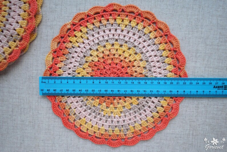 Puede incluir: Un tapete redondo de ganchillo hecho a mano en tonos naranja, amarillo y gris, con un borde festoneado. Una regla azul se coloca sobre el tapete, midiendo aproximadamente 23 cm de di&aacute;metro. Otro tapete es parcialmente visible en la esquina superior izquierda.