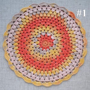 Puede incluir: Un tapete de ganchillo circular hecho a mano con un borde festoneado. El tapete presenta anillos conc&eacute;ntricos de color, incluyendo coral, amarillo y gris. El n&uacute;mero "#1" est&aacute; en la esquina superior derecha.