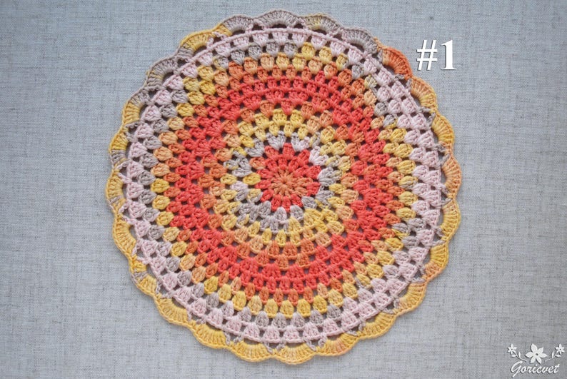 Puede incluir: Un tapete de ganchillo circular hecho a mano con un borde festoneado. El tapete presenta anillos conc&eacute;ntricos de color, incluyendo coral, amarillo y gris. El n&uacute;mero "#1" est&aacute; en la esquina superior derecha.