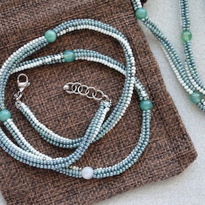 May include: A multi-strand necklace with light blue and white beads and green gemstone accents. The necklace is displayed on a brown fabric surface.