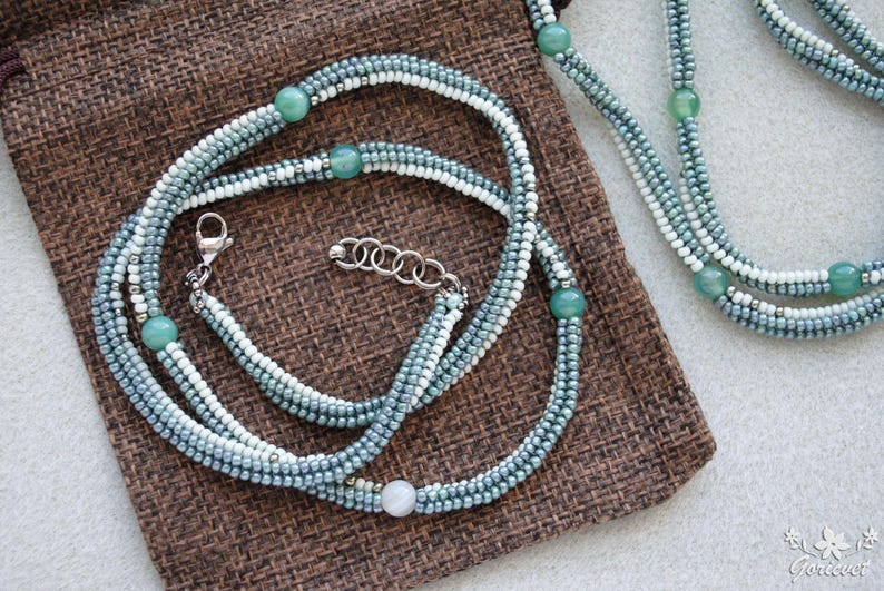 May include: A multi-strand necklace with light blue and white beads and green gemstone accents. The necklace is displayed on a brown fabric surface.