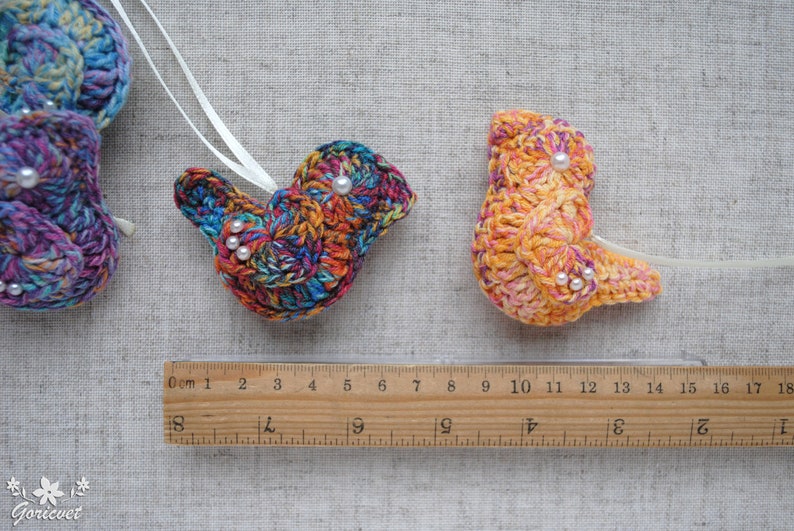 May include: Two crocheted bird ornaments, one with a rainbow pattern and the other with a yellow and orange pattern. Both ornaments have white pearl beads for eyes.