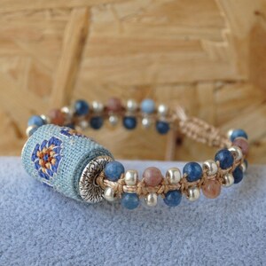 May include: A handmade bracelet featuring a central cylindrical bead wrapped in light blue fabric with a floral design in orange and blue. The bracelet includes blue, pink, and silver beads, strung on a beige cord.