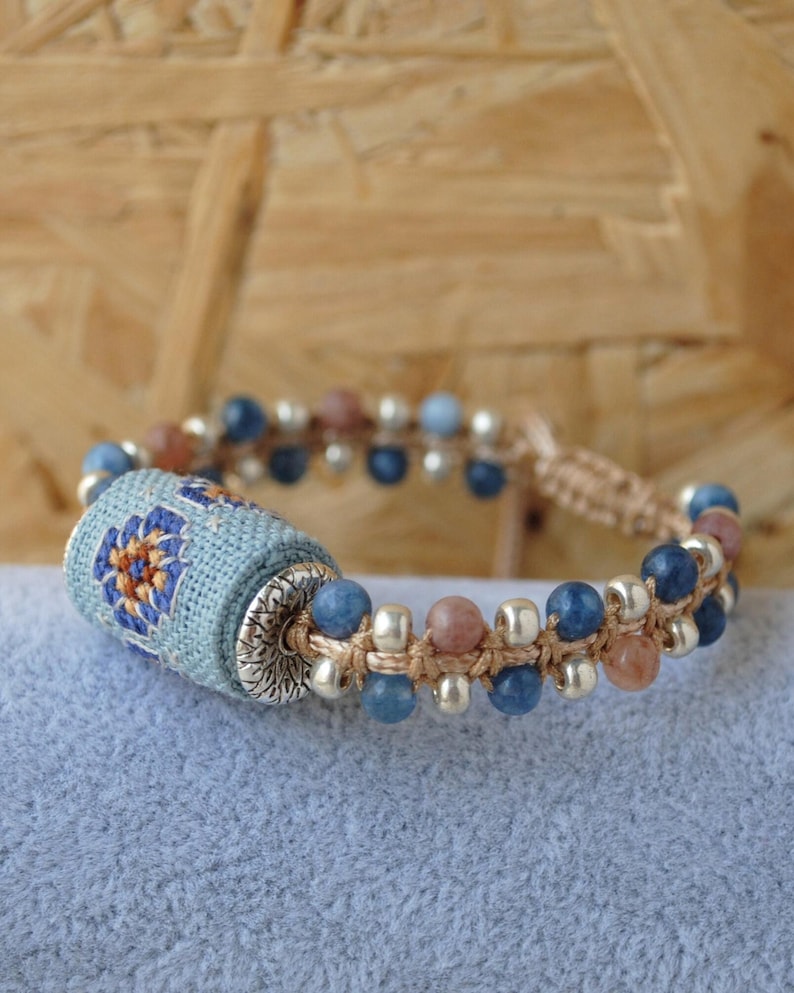 May include: A handmade bracelet featuring a central cylindrical bead wrapped in light blue fabric with a floral design in orange and blue. The bracelet includes blue, pink, and silver beads, strung on a beige cord.