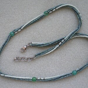 May include: A silver chain necklace with a white and blue beaded strand. The strand is made of small, graduated beads in shades of blue and white. The necklace features three green gemstone beads and one white pearl bead.