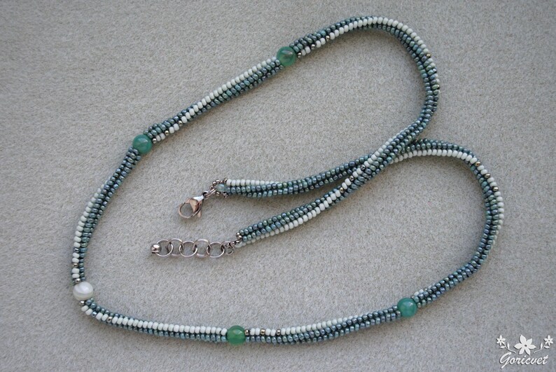 May include: A silver chain necklace with a white and blue beaded strand. The strand is made of small, graduated beads in shades of blue and white. The necklace features three green gemstone beads and one white pearl bead.
