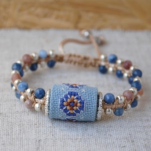May include: Handmade bracelet featuring a central light blue embroidered bead with a geometric design in blue, orange, and brown. The bracelet includes blue, pink, and silver beads, strung on a beige cord.