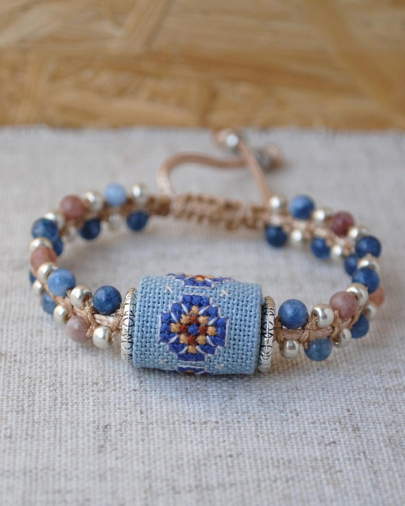 May include: Handmade bracelet featuring a central light blue embroidered bead with a geometric design in blue, orange, and brown. The bracelet includes blue, pink, and silver beads, strung on a beige cord.