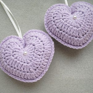 May include: Two lavender crocheted hearts with white pearl accents. The hearts are hanging from white string.