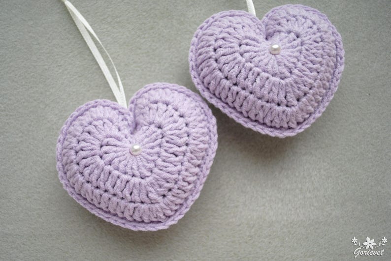 May include: Two lavender crocheted hearts with white pearl accents. The hearts are hanging from white string.