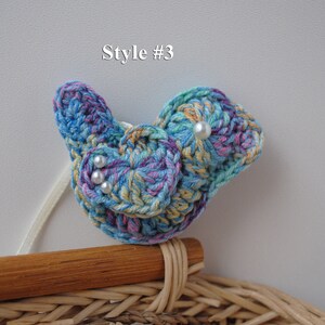 May include: A crocheted blue, yellow, and purple bird with white pearl accents. The bird is sitting on a wooden surface.