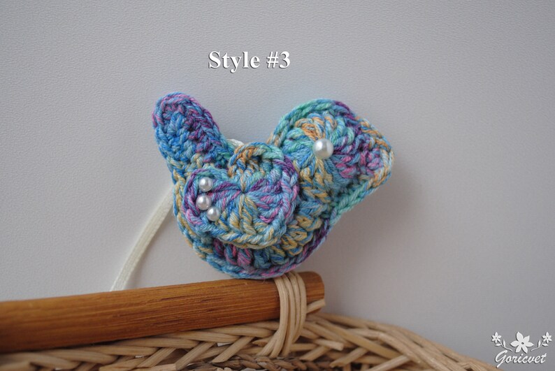 May include: A crocheted blue, yellow, and purple bird with white pearl accents. The bird is sitting on a wooden surface.