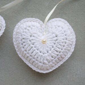 May include: Two white crocheted hearts with a pearl accent in the centre of each heart. The hearts are connected by a white ribbon.