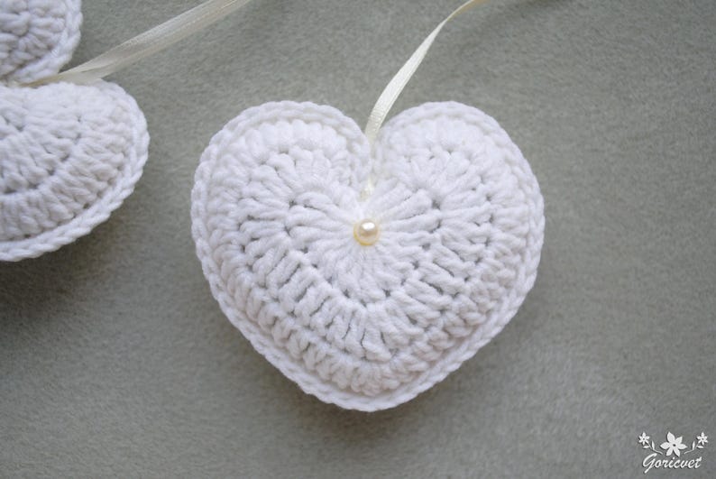 May include: Two white crocheted hearts with a pearl accent in the centre of each heart. The hearts are connected by a white ribbon.
