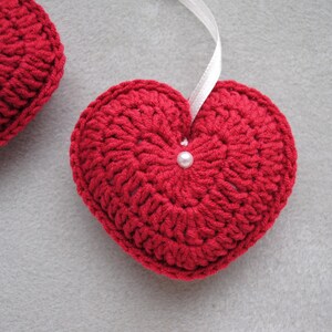 May include: Two red crocheted hearts with white pearl embellishments. The heart on the right is hanging from a white ribbon.