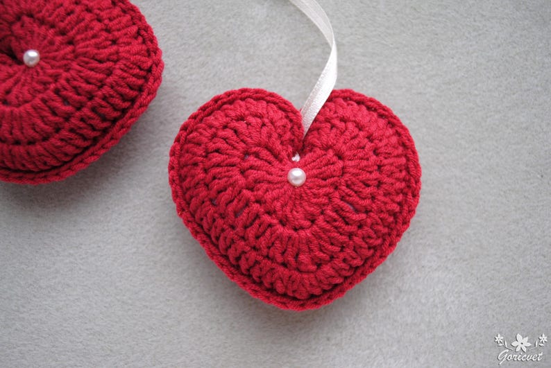 May include: Two red crocheted hearts with white pearl embellishments. The heart on the right is hanging from a white ribbon.