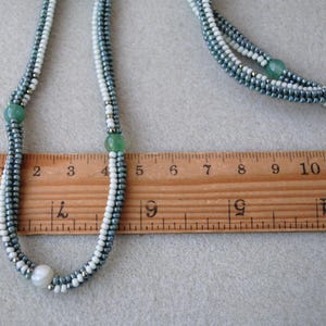 May include: A multi-strand necklace with white and gray seed beads and green gemstone accents. The necklace is measured against a ruler, showing it is approximately 9 inches long.