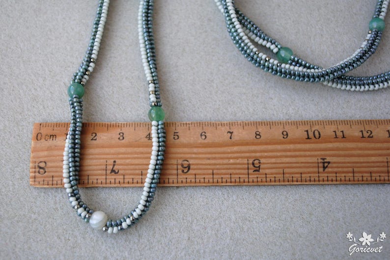 May include: A multi-strand necklace with white and gray seed beads and green gemstone accents. The necklace is measured against a ruler, showing it is approximately 9 inches long.