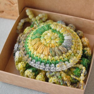 May include: A crocheted flower with green, yellow and grey yarn, accented with small, light green beads. The flower is in a brown box.