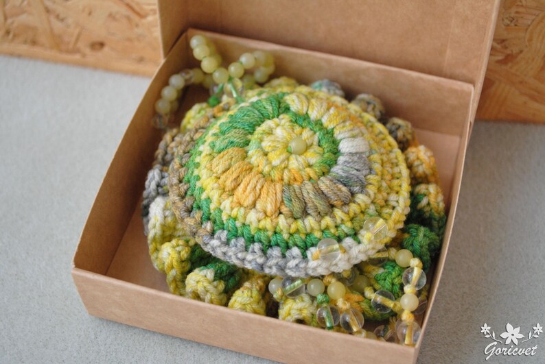 May include: A crocheted flower with green, yellow and grey yarn, accented with small, light green beads. The flower is in a brown box.