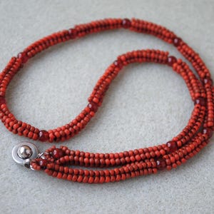 May include: A handmade necklace featuring a triple strand design. The necklace is composed of small, round, red beads, with larger, darker red beads interspersed throughout. A silver-colored clasp secures the necklace.