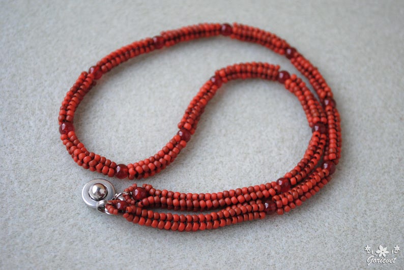 May include: A handmade necklace featuring a triple strand design. The necklace is composed of small, round, red beads, with larger, darker red beads interspersed throughout. A silver-colored clasp secures the necklace.