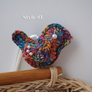 May include: A colorful crocheted bird brooch with white pearl accents. The brooch is attached to a wooden stick and is sitting on a wicker basket.