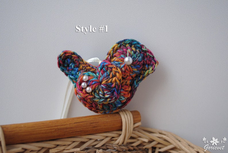 May include: A colorful crocheted bird brooch with white pearl accents. The brooch is attached to a wooden stick and is sitting on a wicker basket.