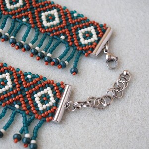 K&ouml;nnte beinhalten: Nahaufnahme eines Armbands mit Perlen und einem geometrischen Muster. Das Armband hat t&uuml;rkisfarbene, rostfarbene und wei&szlig;e Perlen mit h&auml;ngenden t&uuml;rkisfarbenen Perlenakzenten. Der Verschluss und die Kette sind silberfarben.