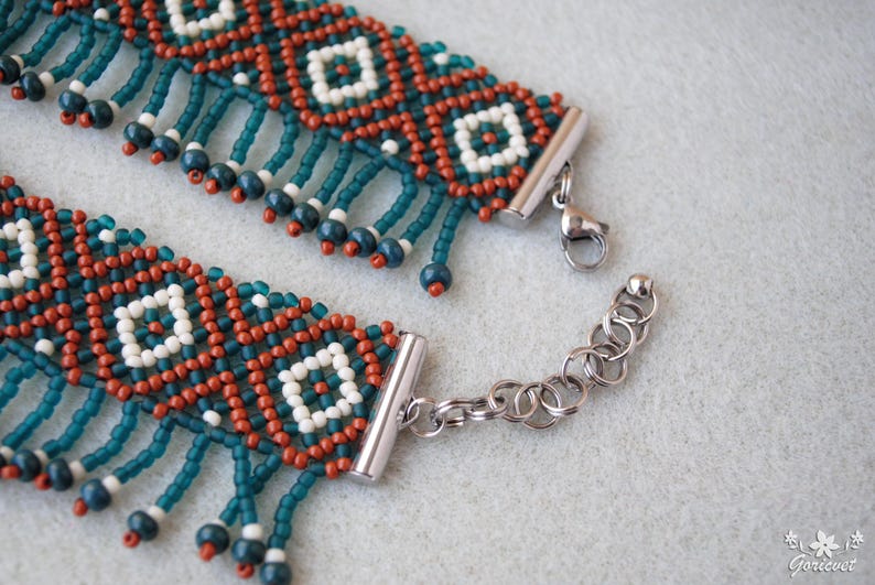 K&ouml;nnte beinhalten: Nahaufnahme eines Armbands mit Perlen und einem geometrischen Muster. Das Armband hat t&uuml;rkisfarbene, rostfarbene und wei&szlig;e Perlen mit h&auml;ngenden t&uuml;rkisfarbenen Perlenakzenten. Der Verschluss und die Kette sind silberfarben.