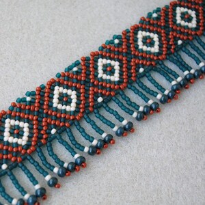 K&ouml;nnte beinhalten: Eine Perlenkette mit geometrischem Muster. Die Kette zeigt ein sich wiederholendes Design aus wei&szlig;en Quadraten in roten und teal-farbenen Rauten. T&uuml;rkisfarbene Perlen h&auml;ngen am unteren Rand, mit wei&szlig;en und marinefarbenen Akzenten.