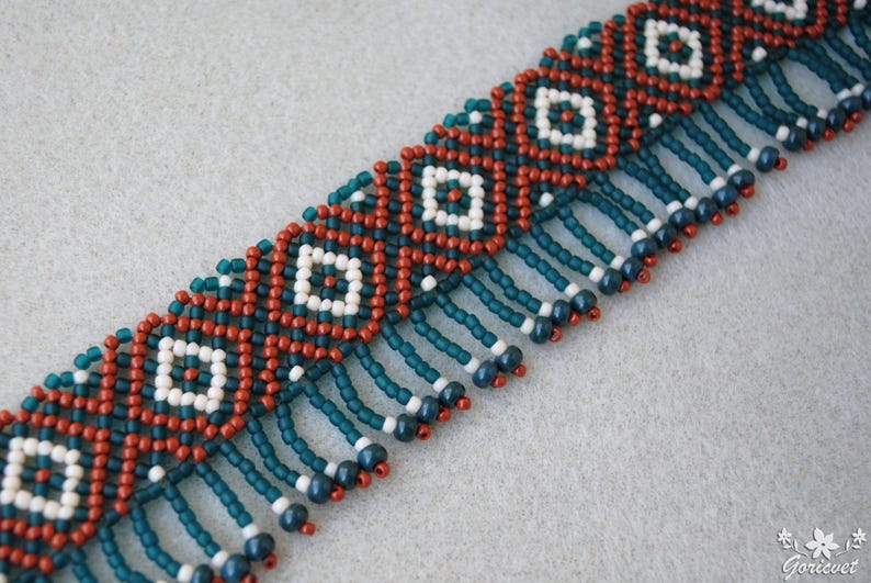 K&ouml;nnte beinhalten: Eine Perlenkette mit geometrischem Muster. Die Kette zeigt ein sich wiederholendes Design aus wei&szlig;en Quadraten in roten und teal-farbenen Rauten. T&uuml;rkisfarbene Perlen h&auml;ngen am unteren Rand, mit wei&szlig;en und marinefarbenen Akzenten.