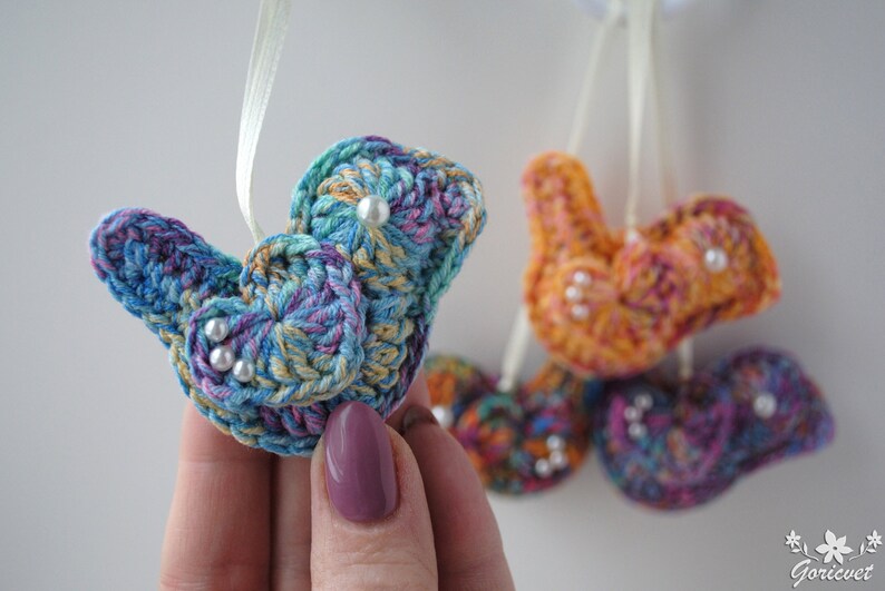 May include: Five crocheted bird ornaments with pearl accents. The birds are in shades of blue, purple, orange, and pink. They are hanging from white ribbon.