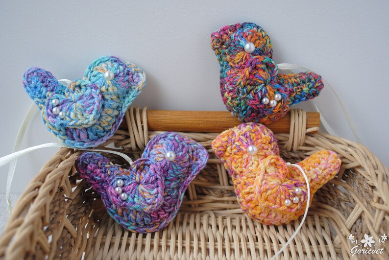 May include: Four crocheted bird ornaments in a wicker basket. The birds are in shades of blue, purple, orange, and green with white pearl accents.