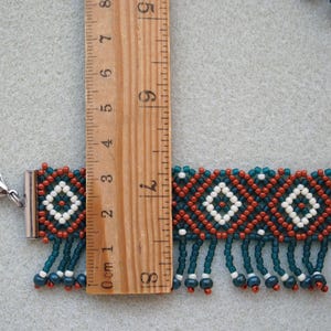 K&ouml;nnte beinhalten: Handgefertigtes Perlenarmband mit geometrischem Muster in Teal, Rost und Wei&szlig;. Das Armband ist ca. 7.6 cm lang und hat einen Silberverschluss und h&auml;ngende Perlen. Das Design umfasst Rauten und Fransen.