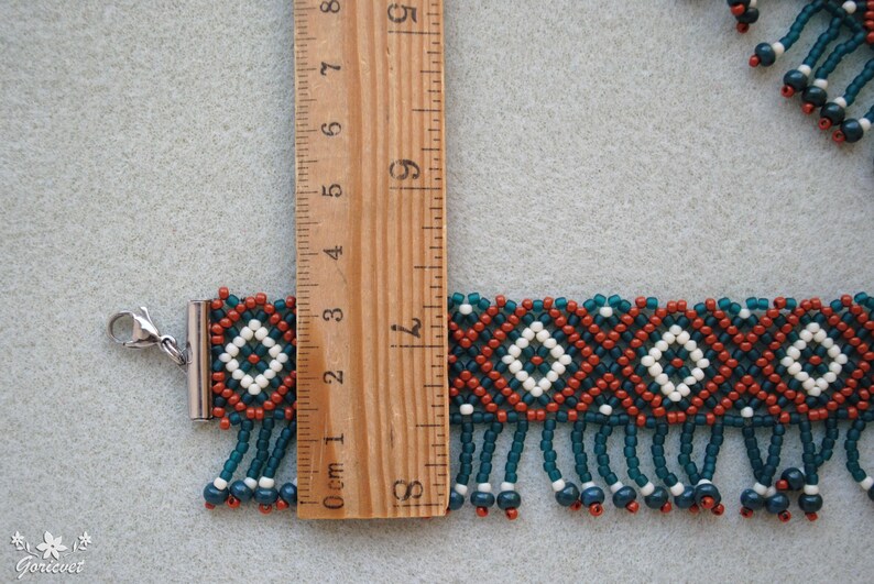 K&ouml;nnte beinhalten: Handgefertigtes Perlenarmband mit geometrischem Muster in Teal, Rost und Wei&szlig;. Das Armband ist ca. 7.6 cm lang und hat einen Silberverschluss und h&auml;ngende Perlen. Das Design umfasst Rauten und Fransen.