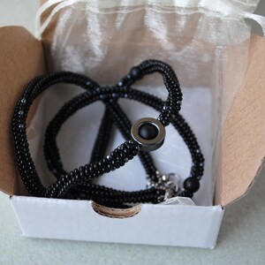 May include: A black beaded necklace with a silver clasp and a black stone pendant. The necklace is displayed in a white box with a white tissue paper lining.