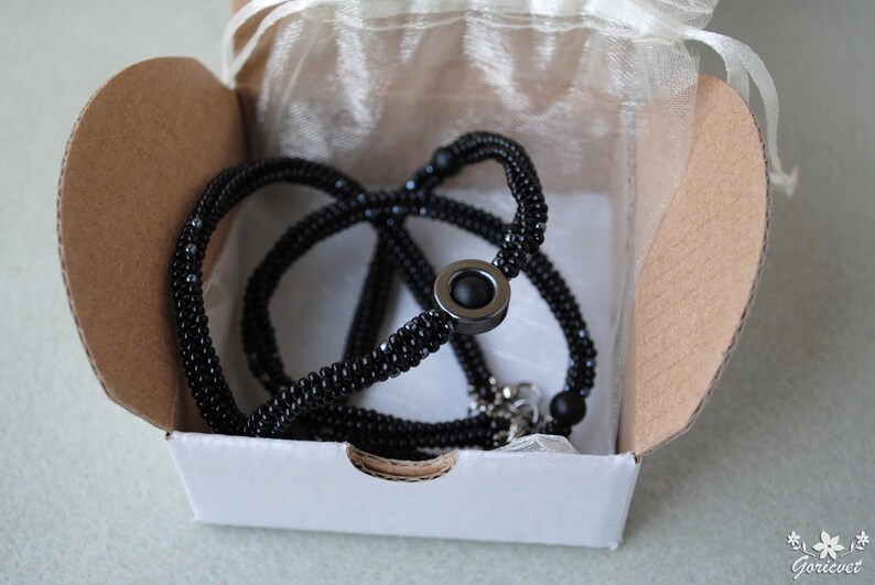 May include: A black beaded necklace with a silver clasp and a black stone pendant. The necklace is displayed in a white box with a white tissue paper lining.