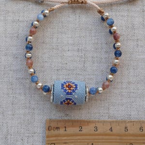 May include: Adjustable bracelet featuring a central light blue cylinder with a floral cross-stitch design. The bracelet includes blue, pink, and silver beads, and is strung on a beige cord. The bracelet is a handmade jewelry item.