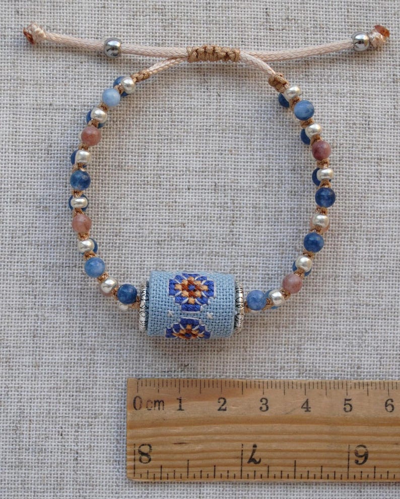 May include: Adjustable bracelet featuring a central light blue cylinder with a floral cross-stitch design. The bracelet includes blue, pink, and silver beads, and is strung on a beige cord. The bracelet is a handmade jewelry item.