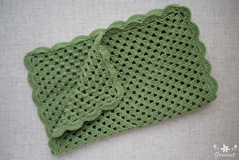 May include: A green crocheted blanket with a scalloped edge. The blanket is folded in half and is lying on a light gray surface.