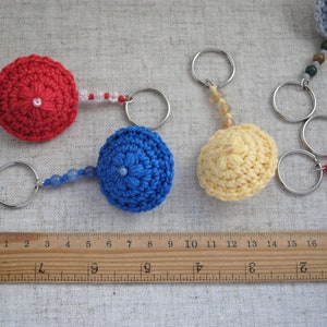 May include: Five crocheted keychains with different coloured beads and metal rings. The keychains are in the shape of small hats, with colours including red, blue, yellow, and grey.