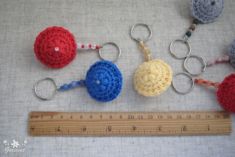 May include: Five crocheted keychains with different coloured beads and metal rings. The keychains are in the shape of small hats, with colours including red, blue, yellow, and grey.