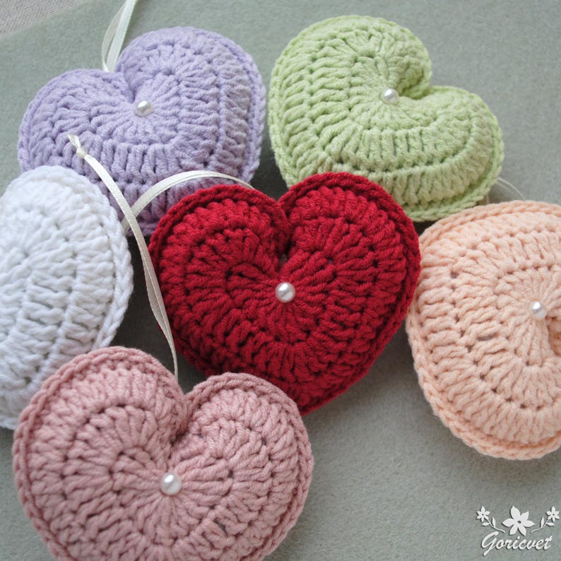 May include: Six crocheted heart-shaped ornaments in various colours, including white, lavender, green, red, and pink. Each heart has a pearl button in the centre.
